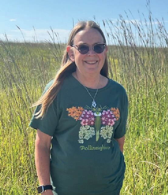 Carol Smith smiling in a butterfly T-shirt in front of tall prairie grass