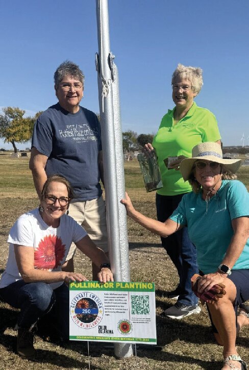 Four women holding a planting sign beside a flagpole at Veterans Park